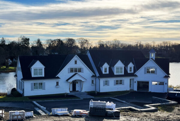 A large, two-story white house with a black roof is set against a lake backdrop. The home features multiple gables and dormer windows. In the foreground, construction materials are visible near a paved driveway. The sky is partly cloudy, and bare trees line the horizon.