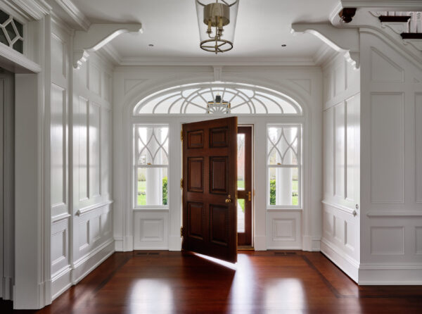 Elegant foyer featuring a richly detailed wooden door partially open, revealing a glimpse of the outside. White-paneled walls and decorative molding frame arched transom windows. A pendant light hangs overhead, and warm wooden flooring enhances the classic design.