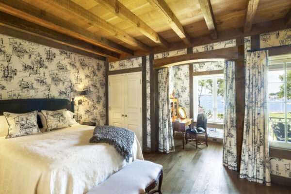 A cozy bedroom features a large bed with a white bedspread and a textured gray throw. The walls are adorned with black and white patterned wallpaper. A wooden ceiling and floor complement the rustic decor. Windows offer a view of trees and a body of water, with a small desk by the window.