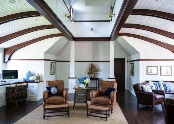 Spacious living room with vaulted white ceilings and wooden beams. Two brown leather armchairs and a sofa surround a coffee table with blue and white flowers. A model sailboat decorates the back. A white desk and chair with a computer screen are on the left. Framed art adorns the walls.