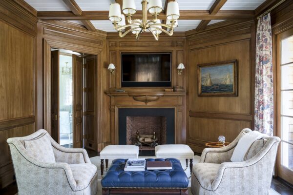 Wood-paneled living room with a central fireplace. Above it, a flat-screen TV is flanked by two sconces. A navy tufted ottoman serves as a coffee table, surrounded by two beige patterned armchairs. A floral curtain hangs next to a window, and a painting of ships adorns the wall.