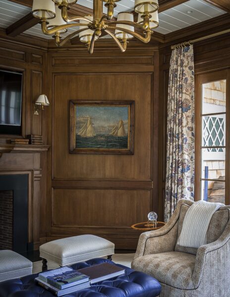 Elegant wood-paneled living room featuring a painting of sailboats, floral curtains, and a decorative chandelier. A cozy armchair, tufted blue ottoman with books, and a fireplace add to the ambiance. A small lamp and a white pouf complete the design.
