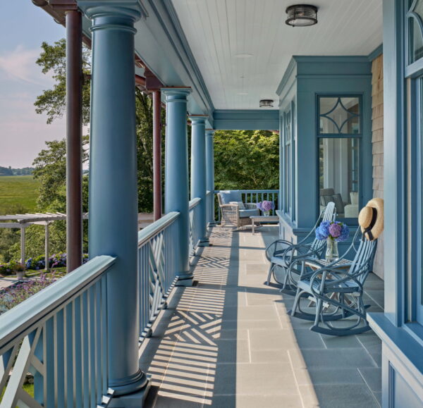 A spacious porch of a shingle-style home features blue wooden columns and railings, with matching rocking chairs. A straw hat rests on one chair as sunlight casts shadows on the stone floor. Lush greenery and a distant landscape enhance the serene and inviting atmosphere.