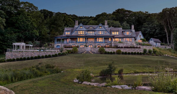 Large, elegant shingle-style country house with intricate gables and expansive windows, surrounded by lush greenery. In front, a manicured lawn with a stone pathway leads to a small pond. Evening light casts a warm glow from the windows, contrasting with the darkening sky and forest backdrop.