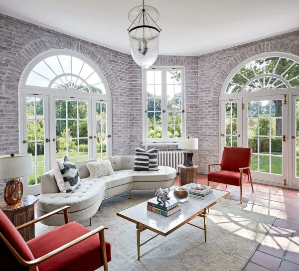 A sunlit sitting room with large arched windows and a view of greenery. It features a curved white sofa with striped cushions, two red armchairs, and a glass-top coffee table on a light rug. A central pendant light hangs from the ceiling. Brick walls add texture to the space.