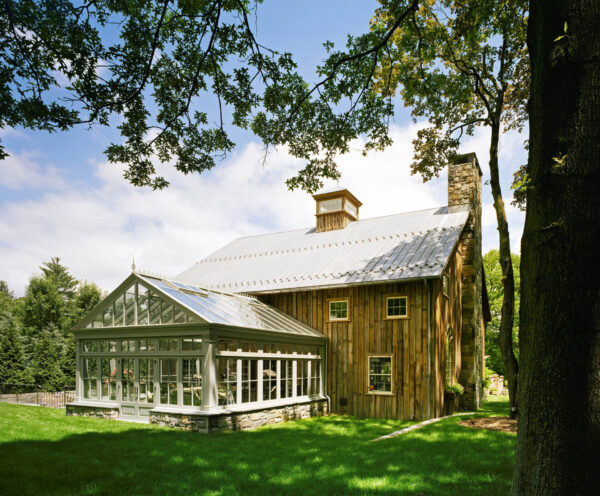 A rustic two-story wooden house with a stone chimney and metal roof is nestled among trees. It features a large glass conservatory on one side. The house is surrounded by lush green grass, with sunlight filtering through the foliage on a clear day.