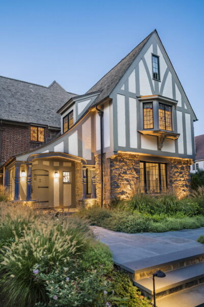 Charming Tudor-style house with stone and timber facade, bay window, and arched entrance exuding classic Tudor architecture vibes. The roof is steeply pitched. A well-maintained garden with grasses and flowers surrounds the stone pathway leading to the entrance. At dusk, warm lights illuminate the exterior.