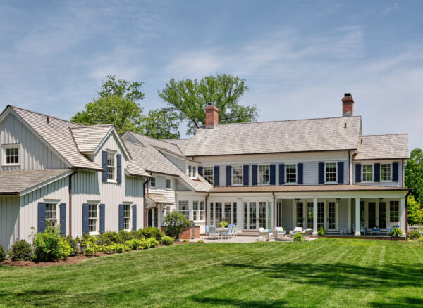 A large, elegant house with white siding and gray-blue shutters is surrounded by a well-manicured lawn. The two-story building features chimneys and a sloped roof. A spacious patio area is furnished with white chairs and tables. Tall trees are visible in the background under a clear blue sky.