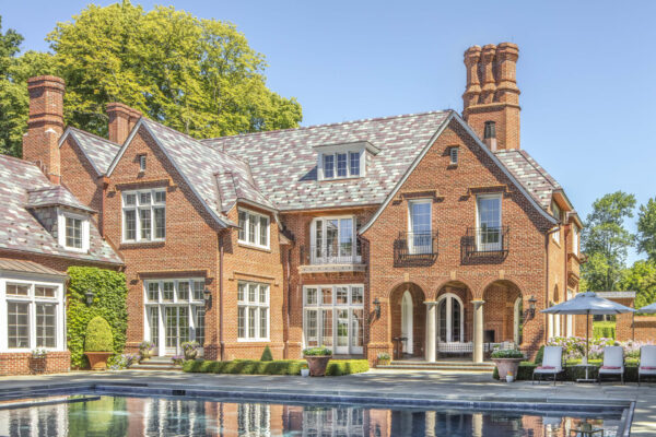 A large, elegant brick mansion with a slate roof and multiple chimneys sits in front of a swimming pool. The house features arched windows and balconies, surrounded by greenery and trees. Poolside chairs with umbrellas are arranged on the stone patio under a clear blue sky.