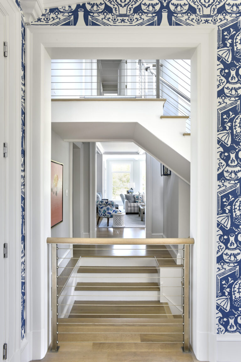 A staircase with light wooden steps and a metal railing leads into a bright, contemporary room. The walls feature blue and white patterned wallpaper. In the background, a seating area with blue-patterned chairs is visible, illuminated by natural light from large windows.