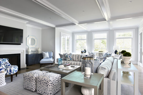 A modern living room with a high ceiling and natural light from large windows. It features a wall-mounted TV above a fireplace, striped sofas, and patterned ottomans. Theres a console table with decorative items and a cabinet topped with vases. The decor is predominantly in grey, blue, and white tones.