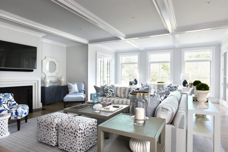 A modern living room with a high ceiling and natural light from large windows. It features a wall-mounted TV above a fireplace, striped sofas, and patterned ottomans. Theres a console table with decorative items and a cabinet topped with vases. The decor is predominantly in grey, blue, and white tones.