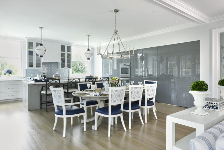 A spacious kitchen and dining area with light wood flooring and white walls. The dining table features white chairs with blue cushions. A large silver chandelier hangs above. The kitchen has a breakfast bar with high stools and modern appliances. Decorative plants and flowers adorn the space.