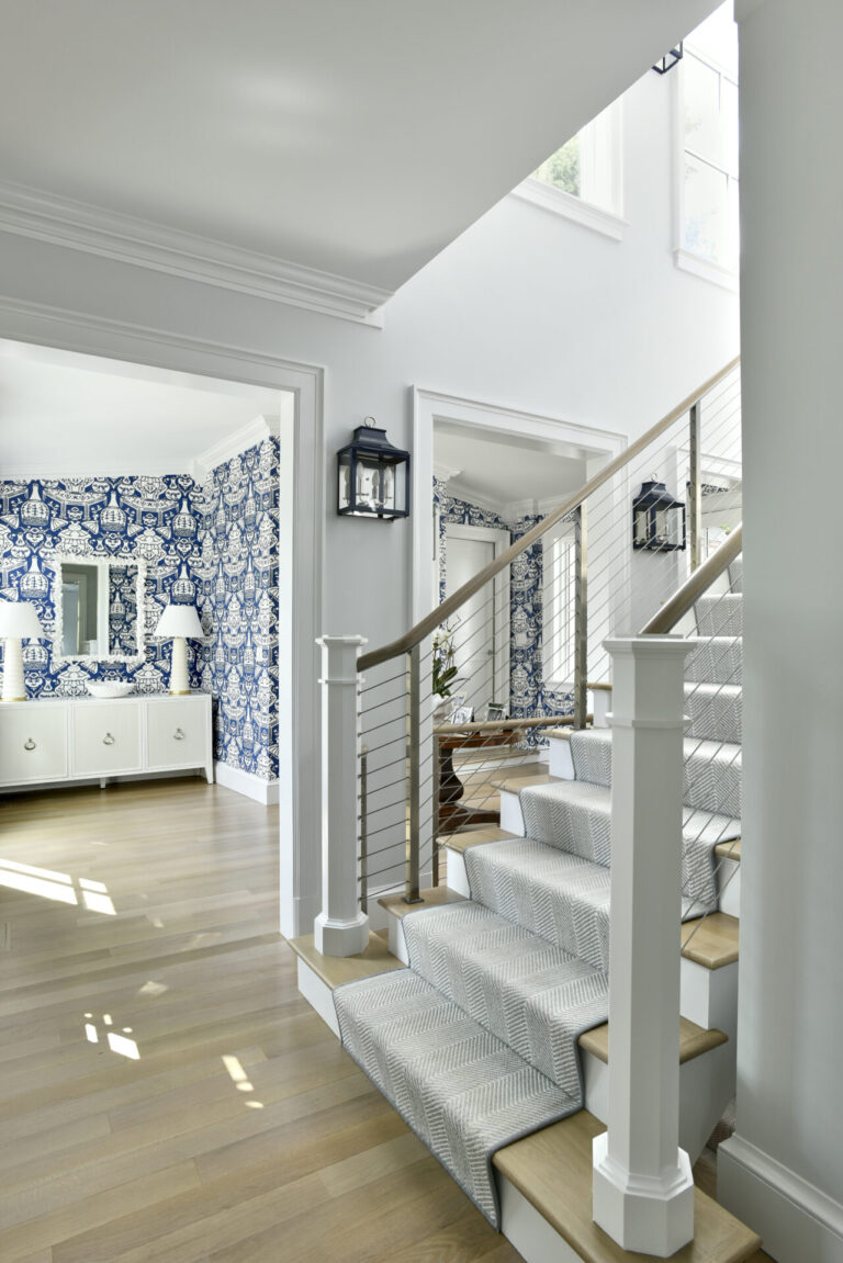 A bright foyer with a staircase featuring a gray runner and white railings. The walls are painted white, and a room with blue and white floral wallpaper is visible. Light hardwood floors and a black wall lantern enhance the airy, modern atmosphere. Natural light floods the space.