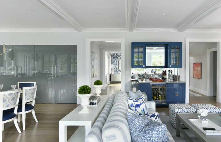 A modern living room with a light gray couch featuring blue patterned cushions. A white table with potted plants sits behind the couch. A navy-blue cabinet/bar area has glassware and bottles. The dining area to the left has white and blue chairs. Bright lighting and wooden floors complete the space.