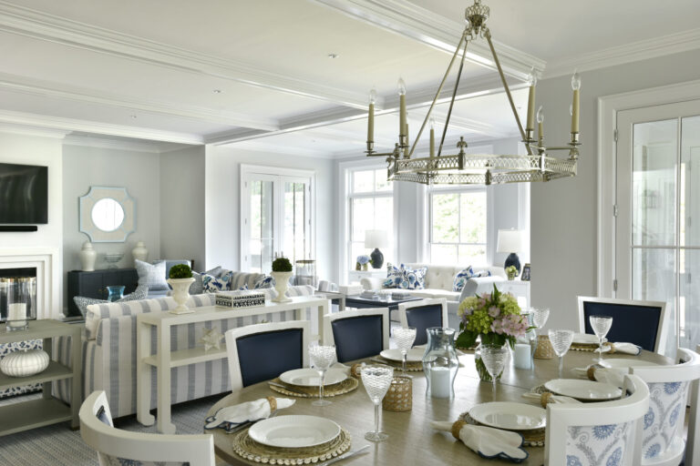 A spacious, elegant living and dining area with a coastal theme. The dining table is set with white plates, wicker placemats, and crystal glassware. A large chandelier hangs above. The living room has white and blue striped furniture, large windows, and a mirror above the fireplace.