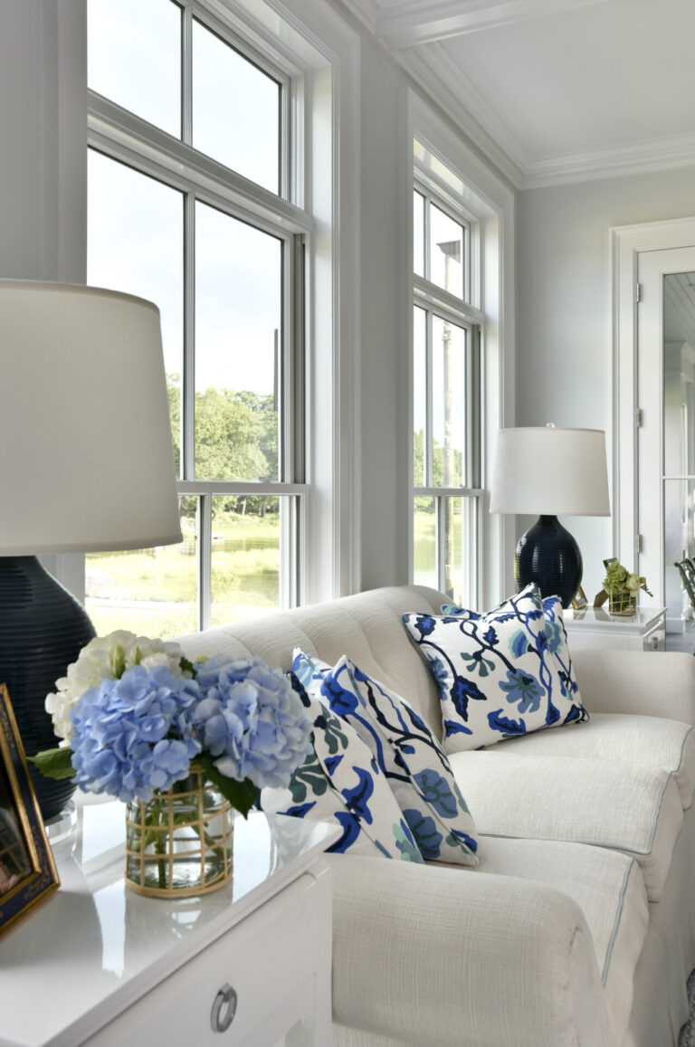 A cozy living room featuring a white sofa adorned with blue and white floral-patterned pillows. A white side table holds a vase of blue hydrangeas and a framed photo. Two large windows with a view of greenery outside and two matching lamps with dark bases complete the serene setting.