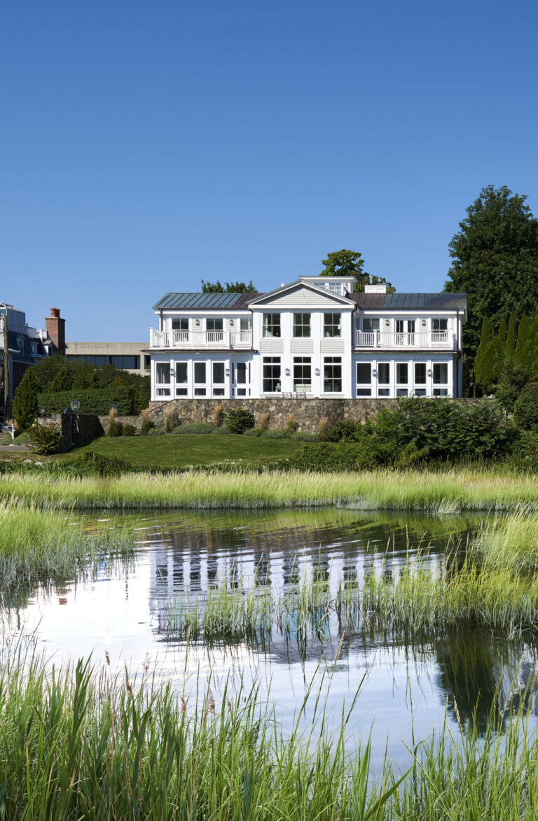 A white two-story house with large windows and a balcony, set on a grassy hillside. Trees surround the house. In the foreground, a reflective pond with tall green grass is visible under a clear blue sky.