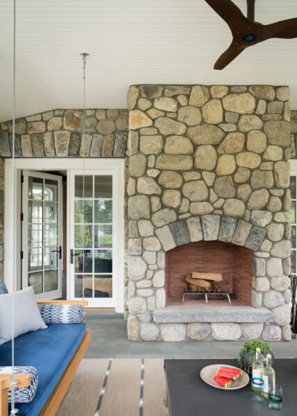 A cozy outdoor room features a large stone fireplace with logs inside. To the left, theres a cushioned swing with blue and gray pillows. A coffee table holds a plant, a plate of watermelon, and a bottle. French doors with glass panes open to a green outdoor view. A ceiling fan is above.
