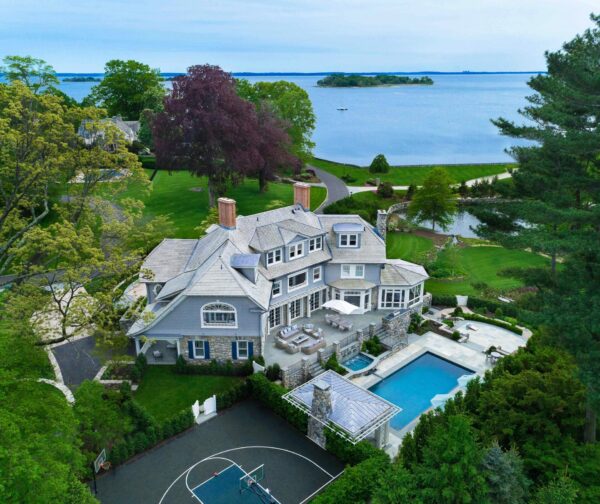 Aerial view of a large, luxurious estate near a body of water. The house is surrounded by lush greenery and features multiple gabled roofs. In the backyard, there’s a swimming pool, patio area, and a basketball court. A winding road and trees lead down to the waterfront, with a small island visible.