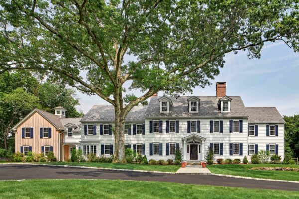 Large colonial-style house with white siding, black shutters, and multiple chimneys. A tall, leafy tree stands prominently in the foreground. The house features a main entrance with columns and a smaller attached building. The lawn is well-kept, and a paved driveway curves in front.