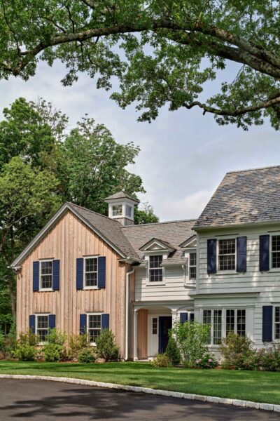 A two-story house features a combination of wood siding and white paint with blue shutters. The left section has vertical wood panels, while the right is painted white. A cupola sits atop the roof. Greenery surrounds the house, and an arched tree branch frames the top of the image.