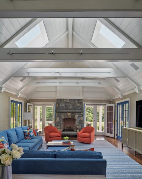 Spacious living room with vaulted ceiling and exposed beams. Features a stone fireplace with a wood mantel. Blue sofas and orange armchairs sit around a low coffee table. Large windows and skylights offer ample natural light. White flowers in a vase on a side table add a fresh touch.