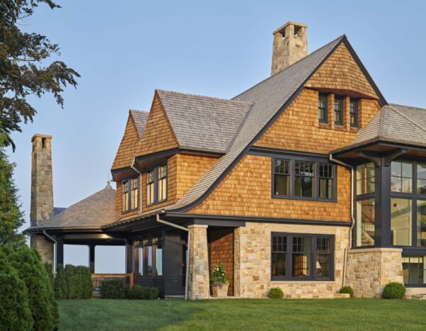 A large, modern house with brown wooden shingles, tall chimneys, and multiple windows. The structure features stone and wood elements, with a wrap-around porch. Its set in a grassy environment with some shrubbery, and the sky is clear and blue, suggesting a sunny day.