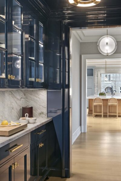 A sleek kitchen with dark blue glossy cabinets and a light marble countertop. On the counter are a cutting board with a lemon and a white cup. The hallway leads to an open space with light wood flooring, an island with white chairs, a modern circular pendant light, and large windows.