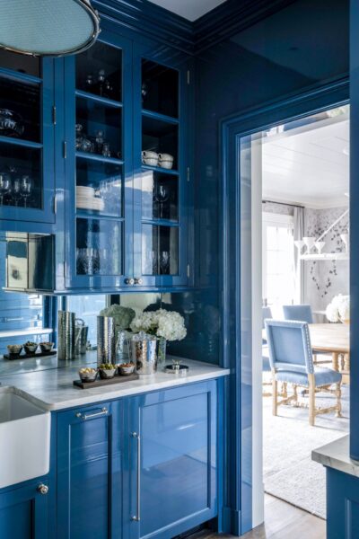 A kitchen with glossy blue cabinets, showcasing glassware and dishes. A white farmhouse sink is visible on the left. Metallic canisters and white hydrangeas in vases are on the countertop. Through the doorway, a dining room with a wooden table and blue-upholstered chairs is seen.