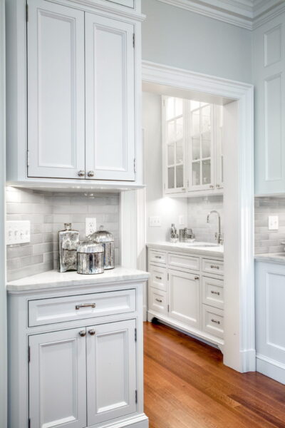 A bright kitchen featuring white cabinets and marble countertops. The backsplash is a glossy gray subway tile. Silver canisters and a lantern adorn the counter. Warm wooden flooring contrasts the white cabinetry, leading to a secondary area with glass-fronted upper cabinets.