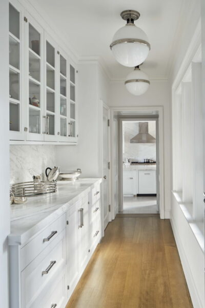 A narrow, bright kitchen hallway with white cabinets and silver handles. Glass-front upper cabinets display dishes. Marble countertops hold utensils. Wooden floor contrasts with the white decor. Two globe pendant lights hang from the ceiling. A view into another room is visible.