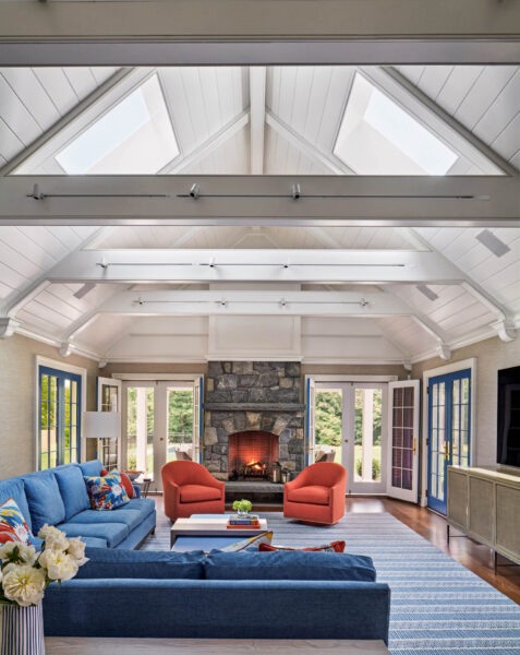Spacious living room with vaulted white ceiling and skylights. Features a stone fireplace with a lit fire, blue sofas, orange armchairs, and a striped rug. Large windows and glass doors offer outdoor views. A bouquet of flowers is on the coffee table, creating a cozy atmosphere.