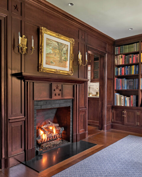 A cozy room features a lit fireplace with a wooden mantel, above which hangs a framed painting. The walls and built-in bookshelves are rich wood, filled with books. Two wall sconces flank the fireplace. The floor is wooden with a gray carpet partially visible.