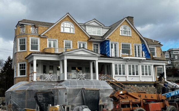 Large two-story house under construction with tan shingles and white trim. Scaffolding, plastic coverings, and construction equipment surround the front. Partial blue tarp covers an area. Cloudy sky overhead, other houses in the background. Stonework visible near entrance.