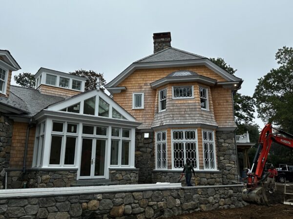 A large house with a mix of stone and shingle siding is under construction. It has a complex roofline with a chimney and multiple windows, including a prominent bay window. A worker stands near an earth mover in the dirt yard, with trees visible in the background under a cloudy sky.