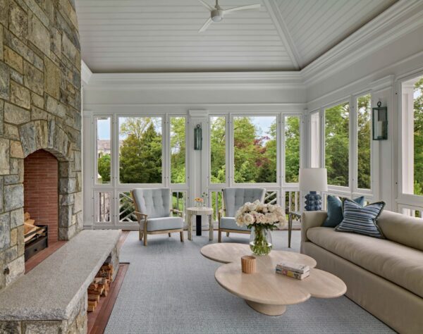 A cozy sunroom with a stone fireplace on the left and large windows showcasing a green outdoor view. It features a beige sofa with cushions, two blue armchairs, and a wooden coffee table with flowers and magazines. The ceiling is vaulted with a ceiling fan, and the floor is carpeted.