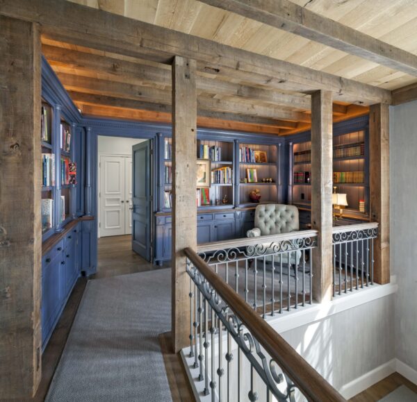 A cozy library with rustic wooden beams and columns overlooks a stairway with wrought iron railings. The room features blue built-in bookshelves filled with books, cabinets, and a tufted gray armchair. Soft lighting highlights the warm and inviting atmosphere.
