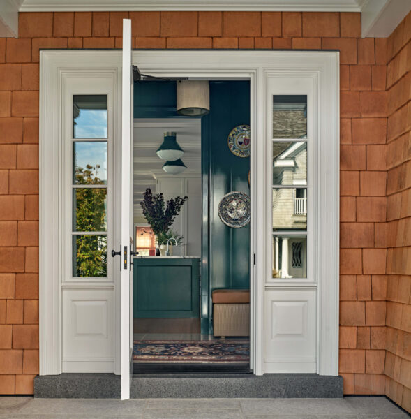 Entrance of a home with white double doors, one slightly open. The exterior features reddish-brown shingles. Inside, a hallway with a wooden floor and a patterned rug leads to a green console table with a vase of purple flowers. Decorative plates and pendant lights adorn the space.