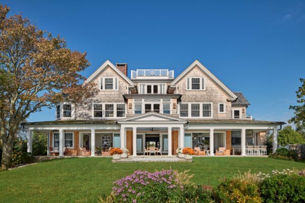 Experience the charm of this large, classic two-story home, part of the Shorefront Shingle Project. With its shingle siding and white trim, gables, dormer windows, and a columned wraparound porch overlooking a vibrant lawn, it invites you to tour an idyllic scene framed by clear blue skies and trees.