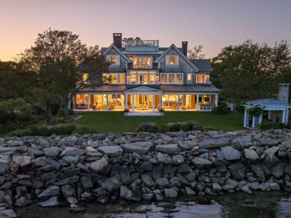 Luxurious three-story house at dusk, illuminated from within, displaying large glass windows. Surrounded by trees, with a stone wall and waterfront in the foreground. Elegant architecture featuring a main building and a connected gazebo, set against a clear evening sky.