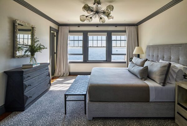 A bedroom with a large window offering a view of the sea. The room features a gray upholstered bed, a gray dresser with a mirror above, a decorative chandelier, and a patterned carpet. Neutral tones and soft lighting create a calming atmosphere. A bench sits at the foot of the bed.