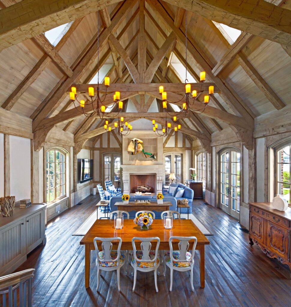 A rustic, vaulted-ceiling living-dining room with exposed wood beams, chandeliers, a wooden dining table, ornate chairs, blue sofas near a stone fireplace, large windows, and wooden floors. A colorful fish sculpture hangs above the mantel. Natural light fills the space.