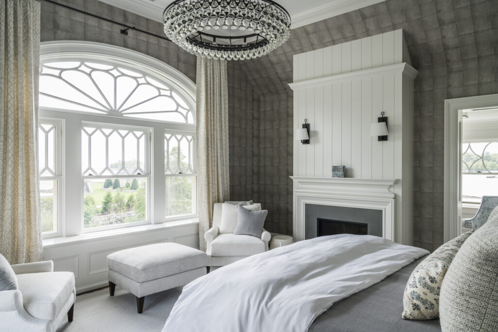 Elegant bedroom with large arched window, two armchairs, and an ottoman. A bed with white linens faces a white paneled fireplace flanked by sconces. A statement chandelier hangs from the ceiling; neutral, soft tones create a serene atmosphere.