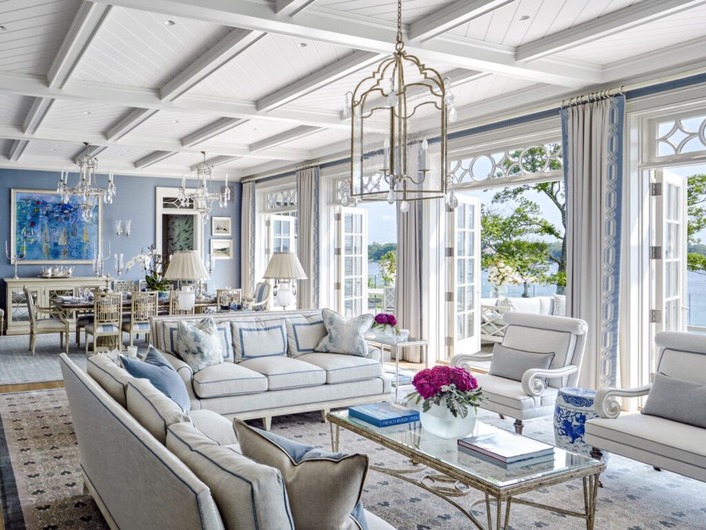 Elegant, airy living room with white sofas, armchairs, and glass coffee tables. Large windows and French doors open to a lake view. Soft blue accents, floral arrangements, and classic decor create a bright, luxurious atmosphere under a coffered ceiling.
