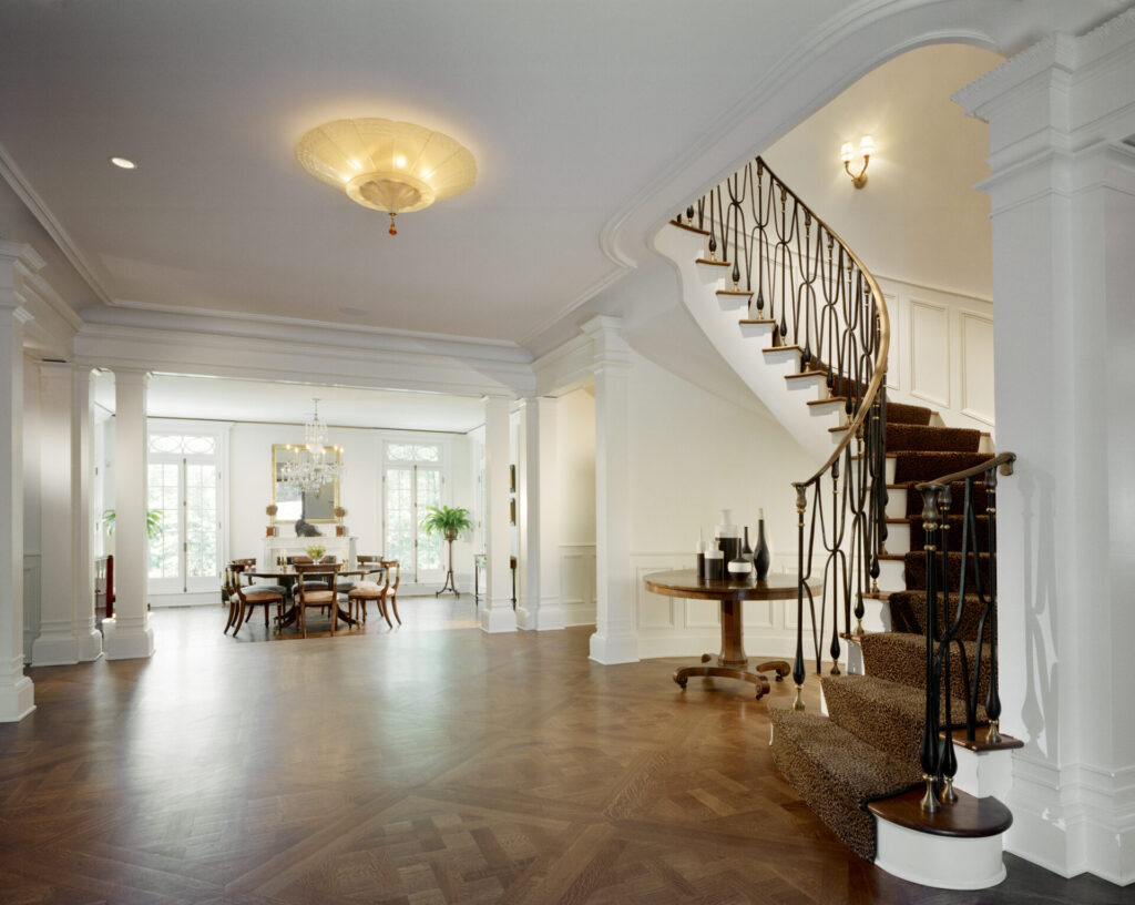 Elegant foyer with polished wood floors, white walls, and an ornate curved staircase with black railings and carpeted steps. A round table with vases sits by the stairs. The space opens into a bright dining room with large windows and a chandelier.