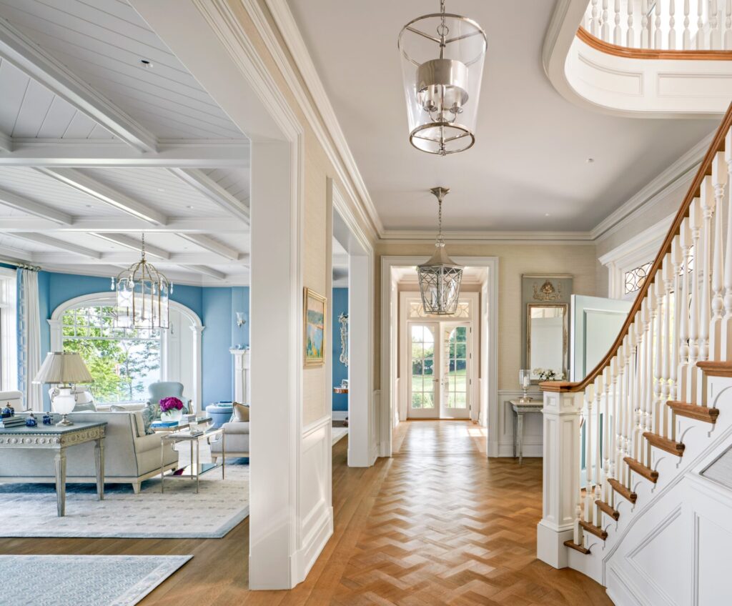 A bright, elegant foyer with parquet wood flooring, a curved staircase with white railings, and a glass lantern chandelier. The space opens to a living room with blue walls, large windows, and classic furnishings in neutral tones. Natural light fills the area.