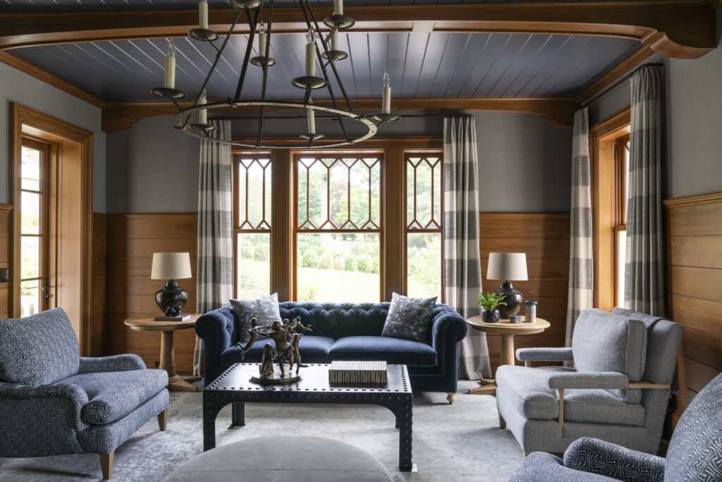 Elegant living room with wood-paneled walls, blue ceiling, and large windows with patterned curtains. Features a dark blue tufted sofa, four blue armchairs, black studded coffee table with a horse sculpture, side tables, and two black lamps.