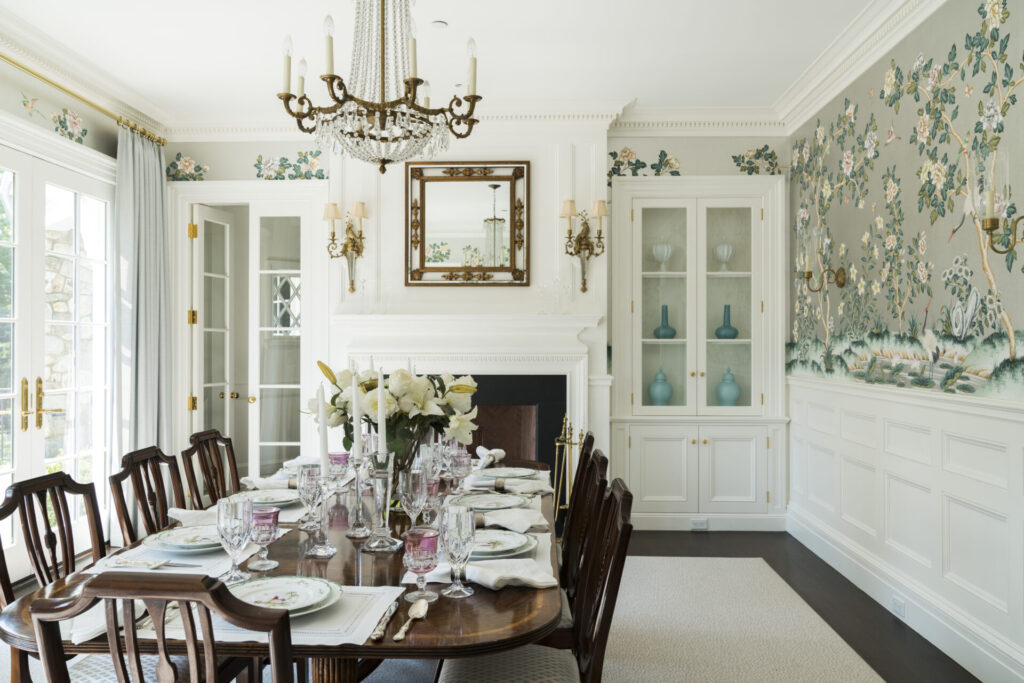 Elegant dining room with a dark wood table set for a meal, white lilies centerpiece, and pastel plates. Floral wallpaper, built-in cabinets with blue vases, a white fireplace, mirror, and chandelier add a classic, bright feel. Large windows let in natural light.