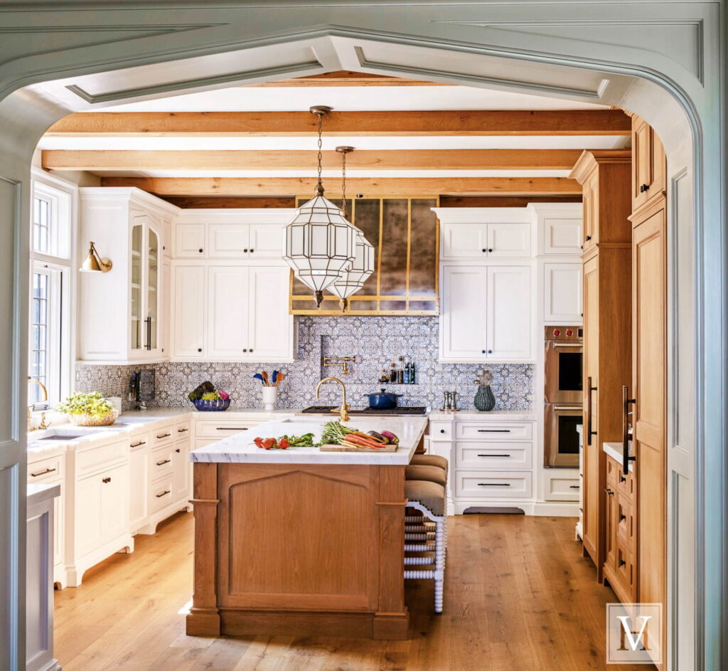 Elegant kitchen with white cabinetry, wood accents, and exposed ceiling beams. Central island with a wooden base and marble countertop is topped with fresh vegetables. Brass hardware, pendant lighting, and a patterned backsplash add stylish details. Sunlight streams in from windows.